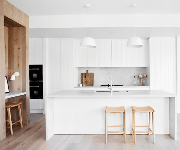 Bright minimalist white kitchen with a large central island, integrated black appliances, vertical-panel cabinetry, pale timber flooring, and warm timber accents. Two pendant lights hang above the island, which includes a sink and seating for two. A timber-lined nook with a small desk and stools is visible to the left.