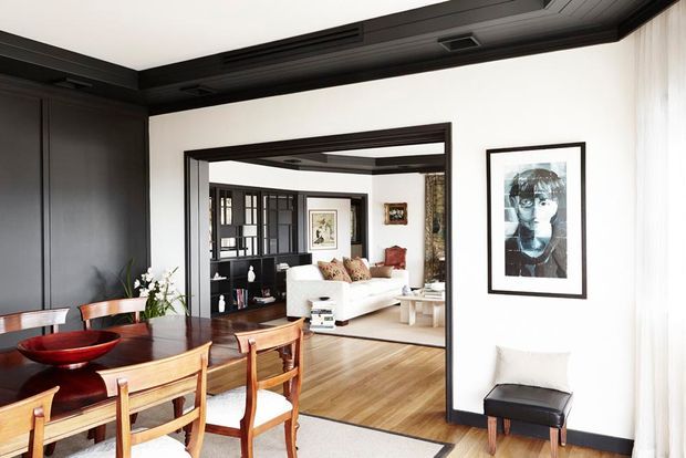 A sophisticated open-plan interior showing a dining area in the foreground connected to a living room beyond. The dining space features a warm timber dining table and chairs, dark built-in wall panelling and cabinetry, and a sculptural red bowl. The adjoining living room has a white sofa, timber flooring, black-framed glazed joinery, artwork, and layered neutral furnishings. Strong black-and-white architectural detailing contrasts with warm wood finishes.