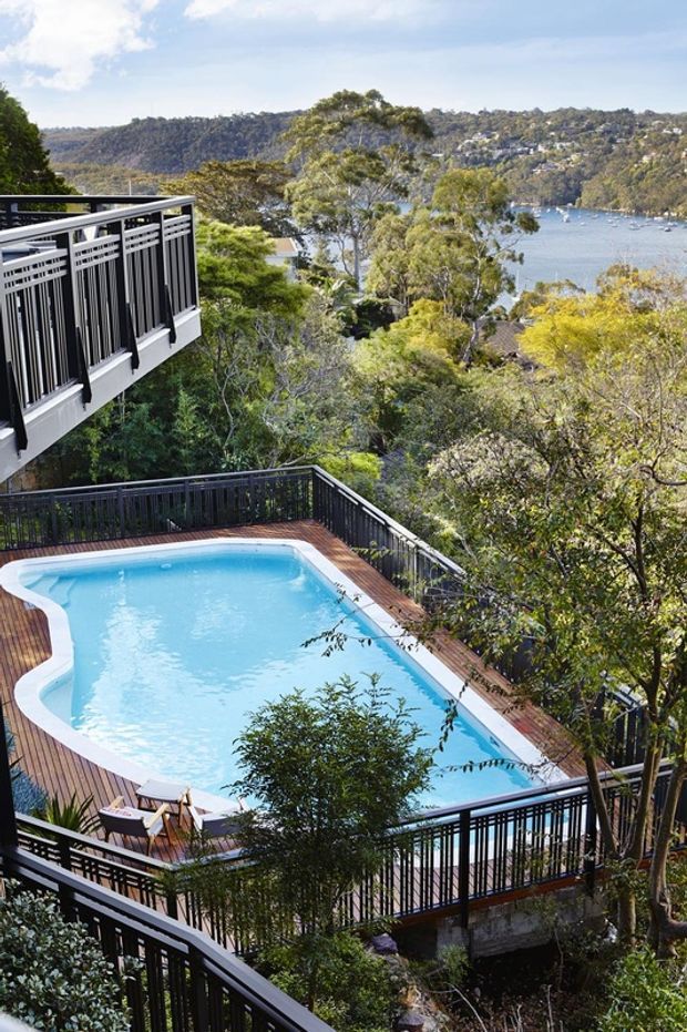 Elevated exterior view of a residential swimming pool surrounded by timber decking and dark metal balustrades, set within a landscaped, wooded site overlooking a broad body of water and densely vegetated hills.