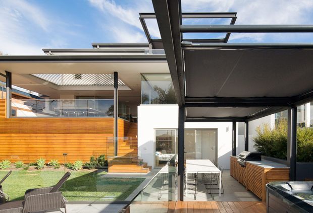 Contemporary residential courtyard and outdoor entertaining area featuring a covered patio, timber slat privacy screen, glass balustrades, built-in timber cabinetry, dining setting, barbecue, and a landscaped lawn. The house has crisp white rendered walls, extensive glazing, slim dark metal framing, and a flat roof with pergola-like overhead structures.