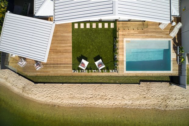 Aerial top-down view of a contemporary residential property with pale horizontal cladding and metal roofing, surrounding a timber deck, central lawn, and rectangular swimming pool. The landscaped outdoor area borders a sandy shoreline or beach-like strip.