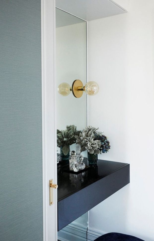 A narrow, refined interior vignette viewed through a partially open door, featuring a large wall mirror, a black floating console with a glossy stone-like top, decorative vases and floral arrangements, and a brass globe wall sconce. The space has white walls and trim, a pale blue textured door, and a polished, softly luxurious aesthetic.