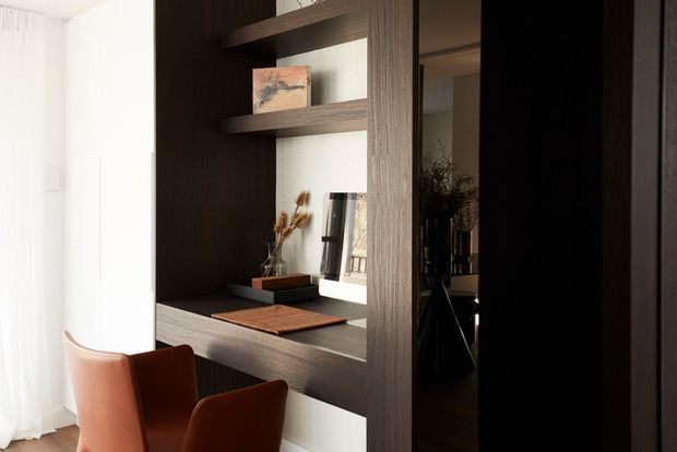 A compact, refined built-in home workspace set within dark wood joinery. The desk features open shelving, a framed artwork, decorative objects, and a mirror or reflective panel, with two warm terracotta upholstered chairs positioned in front. Sheer curtains provide soft natural light from the adjacent window.
