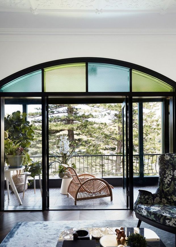 Bright, elegant sunroom or living area opening onto a balcony through a large black-framed arched glass door assembly. The room features a decorative pressed ceiling, dark herringbone timber flooring, patterned upholstered seating, indoor plants, and a woven cane lounge chair. The arched fanlight incorporates translucent pale green and blue glass panels, while mature trees are visible beyond the balcony.