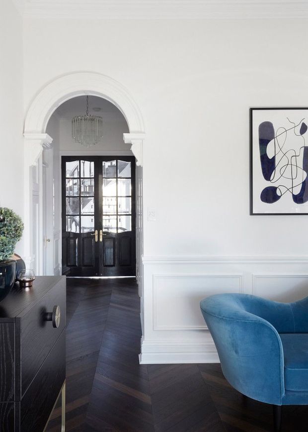 A refined entry hall or living area with white paneled walls, decorative wainscoting, a large arched opening with classical trim, black glazed double entry doors, and dark herringbone timber flooring. A dark built-in or freestanding console sits to the left, while a curved blue velvet armchair occupies the foreground. A framed abstract artwork and hanging crystal light add decorative detail.