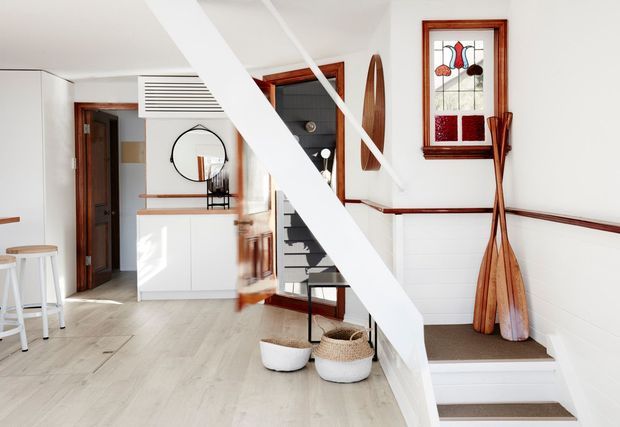 Bright, compact residential entry and stair hall with white walls and cabinetry, pale timber flooring, dark timber trim, and a prominent white structural stair beam. The space includes a small landing, coat or storage cabinetry, a round mirror, open-riser stairs, decorative stained-glass window, and leaning wooden paddles.
