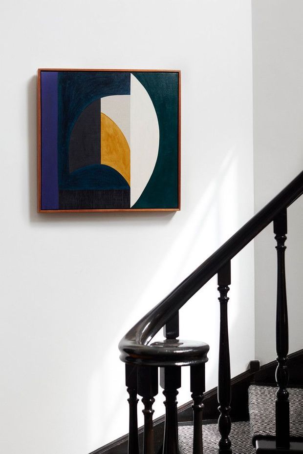 A bright stairway interior with a traditional dark-stained, turned timber banister and balusters, patterned stair runner, and white walls. A framed abstract geometric artwork in deep blue-green, black, mustard, cream, and purple hangs beside the stairs, with sunlight casting a soft highlight on the wall.