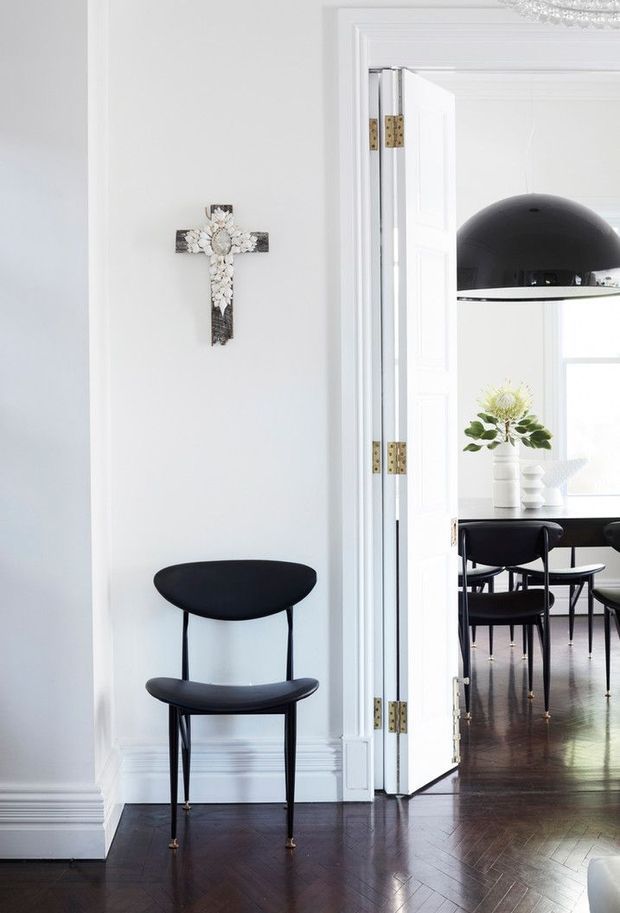 A refined white hallway or entryway with ornate wall mouldings, white paneled double doors, and dark polished timber flooring. A sculptural black chair sits beneath a decorative cross, while the open doors reveal a coordinated dining room with a large black dome pendant, dark dining furniture, and a white floral arrangement.