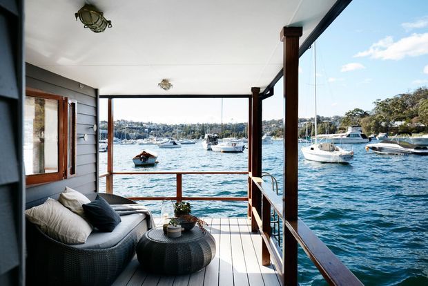 A covered waterfront balcony or deck overlooking a busy harbour, furnished with a woven outdoor lounge chair, cushions, and a round ottoman table. The space has dark weatherboard wall cladding, timber posts and railings, pale timber decking, and extensive clear glazing or screened openings framing boats and water views. The atmosphere is bright, relaxed, and coastal.