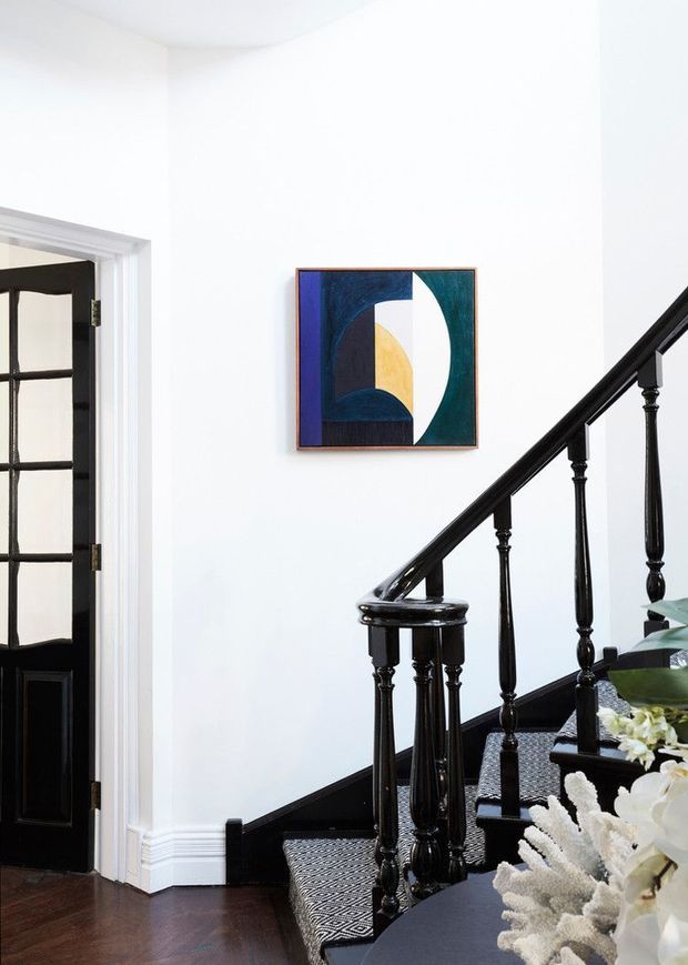 Bright, refined interior view of a traditional stairway and adjacent entry area. The space has white walls with detailed baseboard moulding, dark stained timber flooring, a black-painted timber staircase with turned balusters and a patterned stair runner, and a black glazed-panel door. A colourful abstract artwork hangs on the wall, while floral decor partially occupies the foreground.