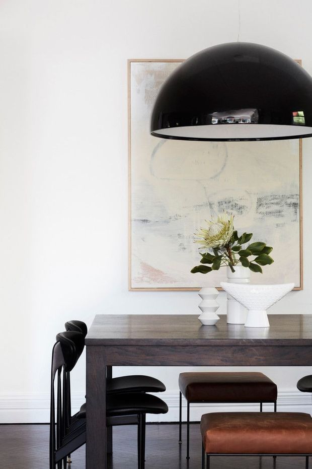 A refined dining area with a dark timber dining table, black sculptural dining chairs, and brown leather stools. A large black dome pendant light hangs above the table, with white ceramic vessels and greenery arranged in front of a pale abstract artwork. The room has white walls, dark wood flooring, and a restrained contemporary aesthetic.