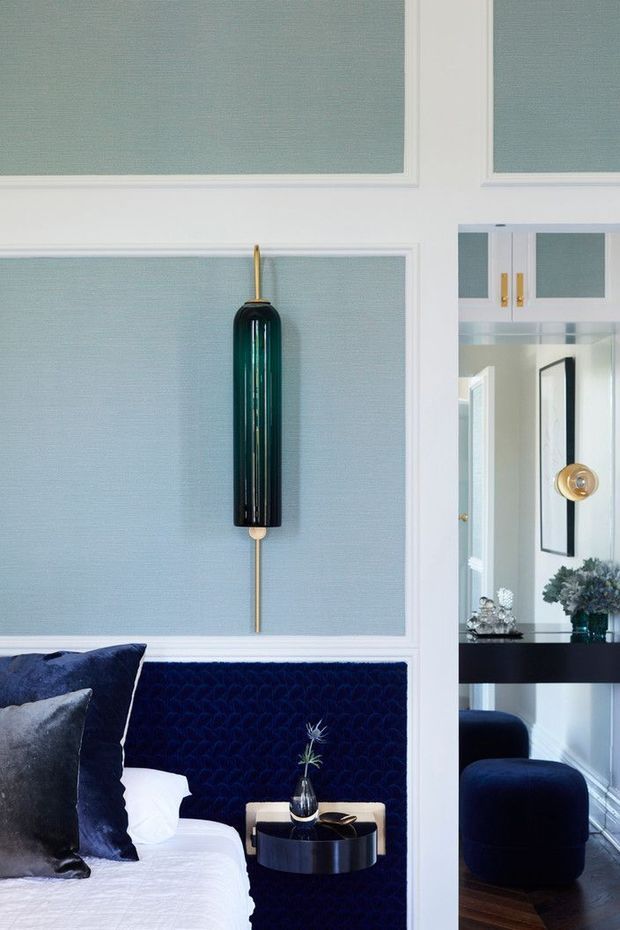 A refined bedroom interior with a deep navy upholstered bedhead and matching bedding accents, set against pale blue-green wall panels framed by white moulding. A sculptural dark glass and brass wall light is mounted above the bed, while a small round black bedside table holds a vase and accessories. A mirrored opening reveals a coordinated adjoining sitting or dressing area with dark blue upholstery, brass detailing, and dark timber flooring.