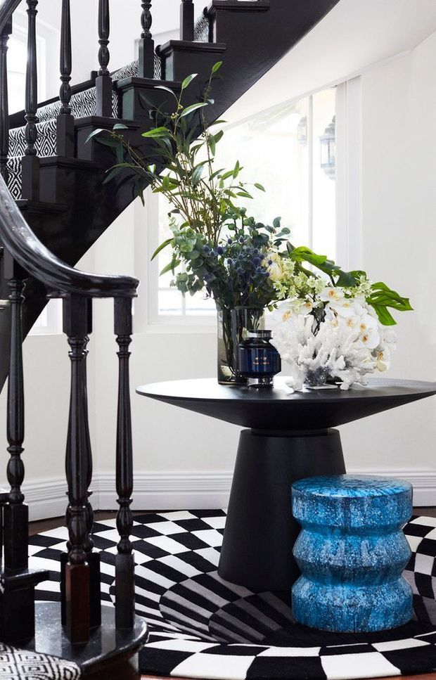 A dramatic black-and-white stair hall with a glossy black staircase and turned balusters, geometric checkerboard flooring, and bright white walls and windows. A sculptural black pedestal table holds large floral arrangements, accompanied by a vivid blue textured stool. The space combines bold monochrome architectural details with colourful decorative styling.