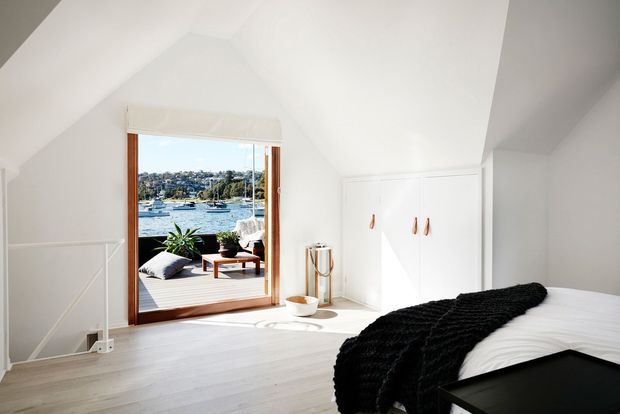 Bright attic-style bedroom with a sloped white ceiling, pale timber flooring, built-in white wardrobe, and a large timber-framed glazed door opening to a small waterfront deck. The room is furnished with a bed dressed in white linen and a black textured throw, with a small black side table and minimal accessories. Boats and a residential waterfront landscape are visible outside.