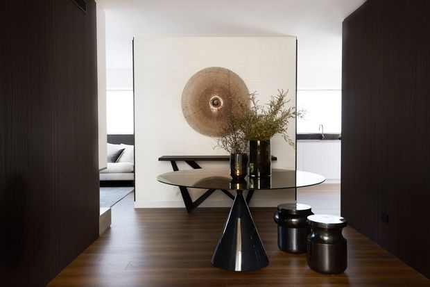 A refined contemporary interior viewed through a dark timber-lined passage, featuring a round dining table with a sculptural black pedestal base, dark cylindrical stools, and a large abstract circular wall artwork. Warm timber flooring, pale walls, black accents, and organic branch arrangements create a sophisticated, minimalist dining setting. A living area and kitchen are partially visible beyond.