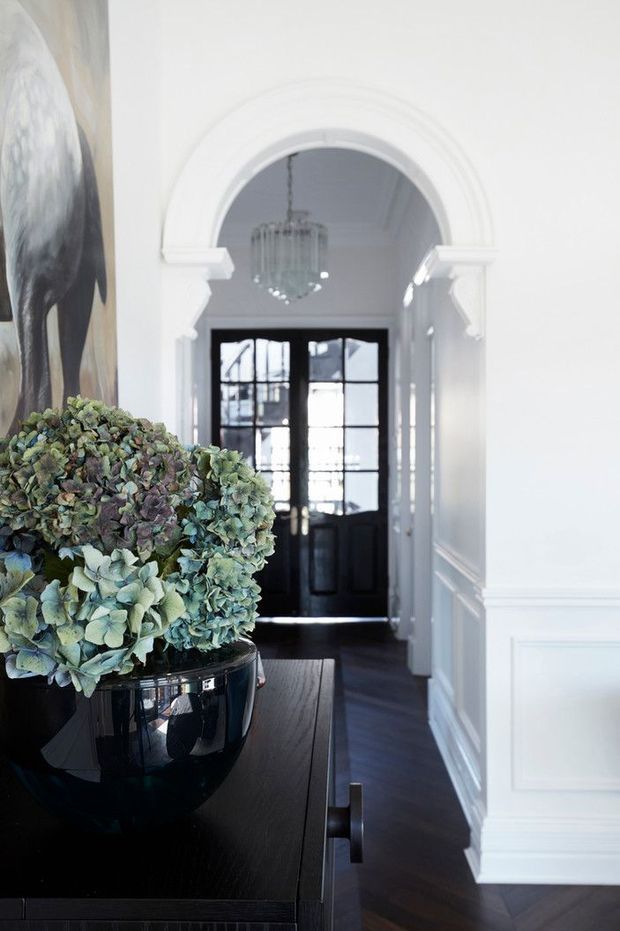 Elegant residential entry hall with a dark timber console and glossy black planter in the foreground, white paneled walls, an ornate arched opening with classical trim, a crystal chandelier, and black glazed double entry doors at the end of the corridor. A large artwork and hydrangea arrangement add decorative detail.