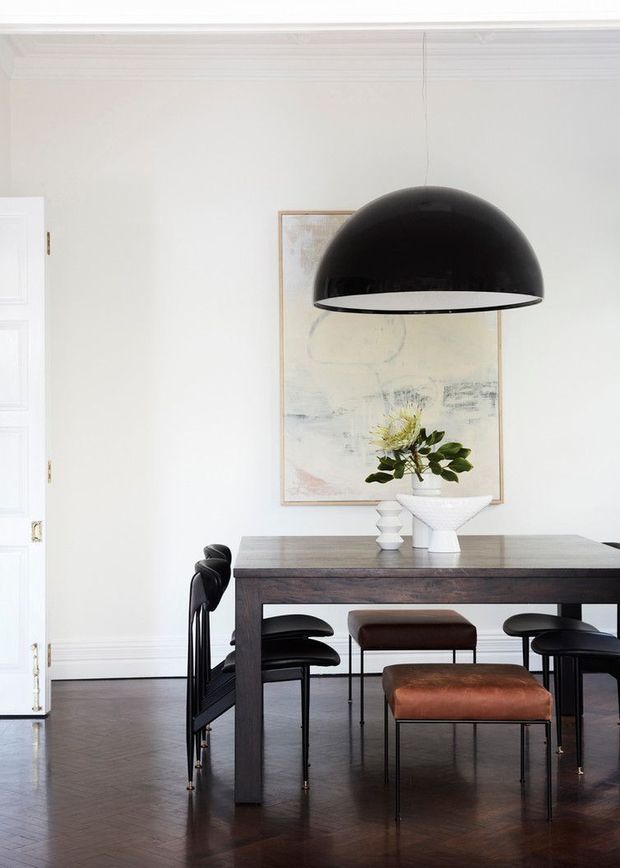 A refined dining room with a dark timber dining table, black and brown upholstered dining chairs, and a large black dome pendant light suspended above the table. The room features white walls with traditional decorative cornice and panelled detailing, dark herringbone timber flooring, a framed abstract artwork, and minimalist floral arrangements.
