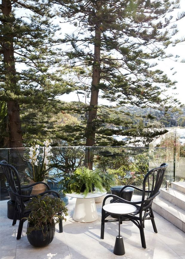 A sunlit outdoor terrace overlooking a leafy coastal or suburban landscape. The terrace has pale tiled paving, frameless glass balustrading, broad steps, black woven chairs, a small white pedestal table, and abundant potted greenery. Tall conifer-like trees dominate the view, creating a private, garden-like setting.