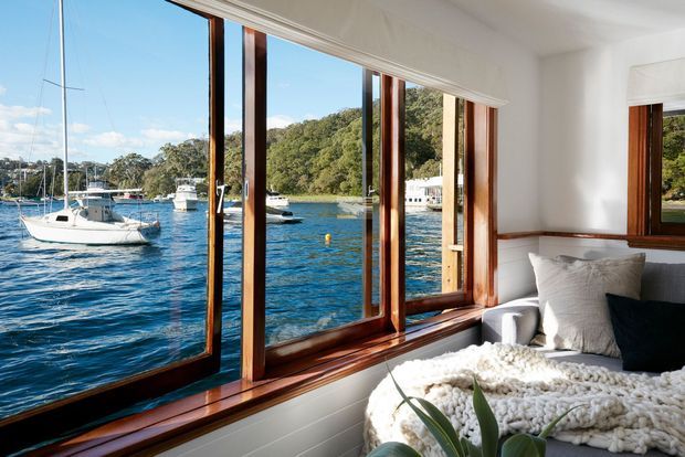 A bright bedroom with a bed positioned beside expansive timber-framed windows overlooking a calm blue-water harbour with boats and wooded shoreline. The room has white walls, warm timber window trim, layered neutral bedding, and strong indoor-outdoor visual connection.