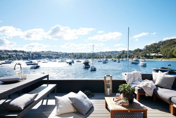 A bright waterfront outdoor terrace overlooking a sheltered harbour, furnished with a timber dining table and bench, a cushioned outdoor lounge, and decorative lanterns and plants. Boats and a leafy shoreline are visible beyond the dark-framed terrace railing.