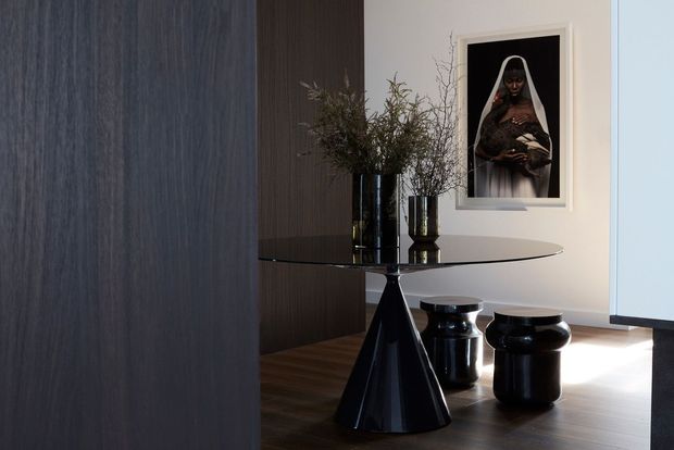 A sophisticated, moody dining area with a glossy black sculptural pedestal table, matching rounded black stools, and dark wood flooring. A dark vertical timber feature wall contrasts with a pale wall displaying framed artwork, while tall glass vases with branches add an organic element. The space has a refined, contemporary minimalist feel with strong black-and-white contrast.