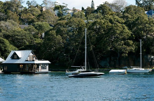 A waterfront scene viewed across blue water, showing a dark-clad floating or waterside house with a steep metal gabled roof and deck, several moored sailboats, and a densely wooded residential shoreline behind it.