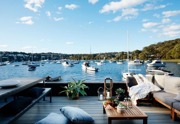 A bright waterfront outdoor lounge on a timber deck overlooking a sheltered marina or harbour. The space includes an L-shaped upholstered sofa with cushions and a throw, a low timber coffee table with potted plants and lanterns, dark outdoor seating, and a small potted palm. Boats, calm blue water, and a tree-covered residential shoreline are visible beyond the deck.