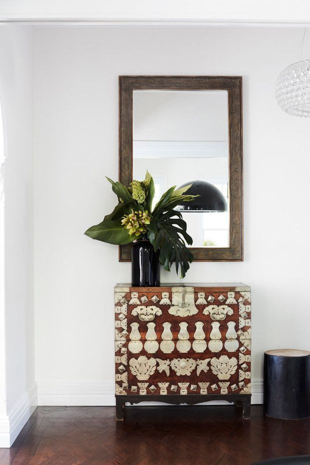 A refined interior vignette featuring a heavily patterned antique-style console cabinet against a white wall, topped with a tall black vase of tropical greenery. A large dark-framed rectangular mirror hangs above, reflecting a black dome pendant light. The space has dark timber parquet flooring, white architectural trims, and a small black cylindrical side table or stool.