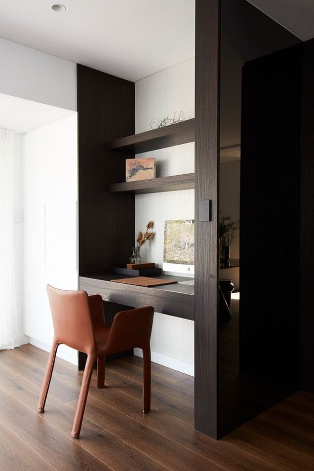 A compact built-in home office nook integrated into a dark timber partition, with a floating desk, open display shelves, and a rust-coloured upholstered chair. The space has white walls and ceiling, dark timber joinery, warm timber flooring, and bright natural light filtering through sheer curtains.