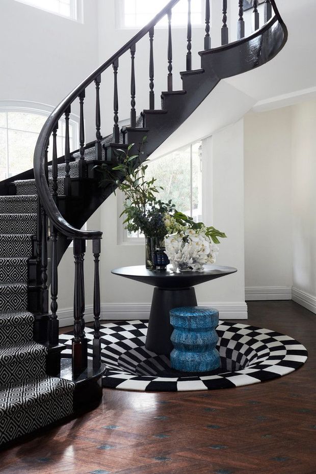A bright, elegant stair hall featuring a dramatic curved black staircase with turned balusters and patterned black-and-white carpeted treads. The space has white walls, large windows, dark polished timber flooring, and a bold black-and-white optical-pattern rug. A black pedestal table with floral arrangements and a vivid blue sculptural stool add decorative contrast.