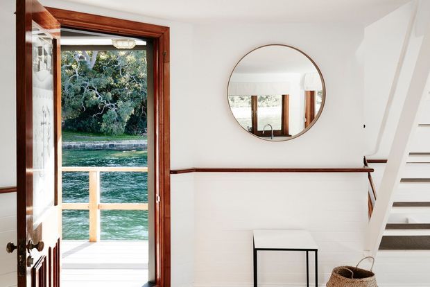Bright interior entryway or stair landing with white painted walls and horizontal wall panelling, dark timber trim, an open timber-framed glazed door leading to a waterside deck, a round wall mirror, a small console table, woven basket, and white staircase.