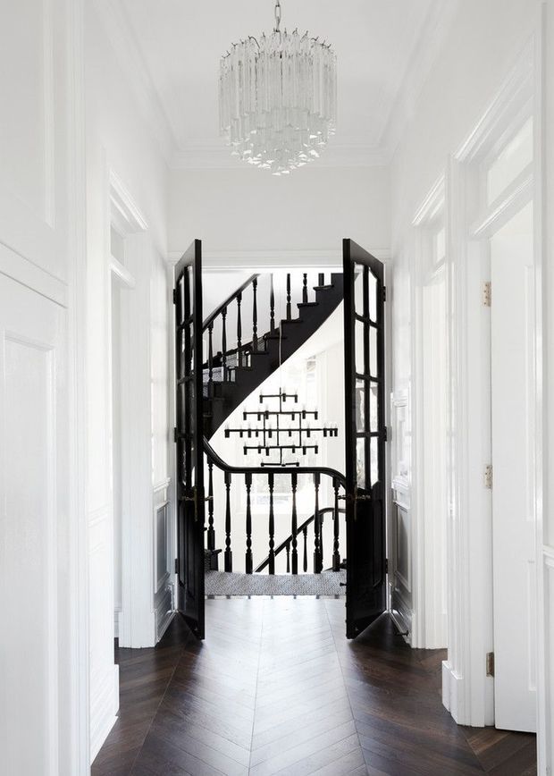 A bright, refined residential hallway leading to an ornate black staircase. The white corridor has detailed wall panelling and trim, tall glazed doors, dark herringbone timber flooring, and a crystal chandelier. Open black-framed double doors frame the stairwell, which features turned balusters and a sculptural curved handrail.