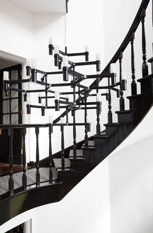 A dramatic interior view of a sweeping black-painted staircase with turned timber balusters and patterned runner carpeting. A striking multi-arm black chandelier with frosted cylindrical bulbs hangs through the stair void, contrasting with crisp white walls and ceilings. Dark timber doors and traditional detailing are visible at the landing.