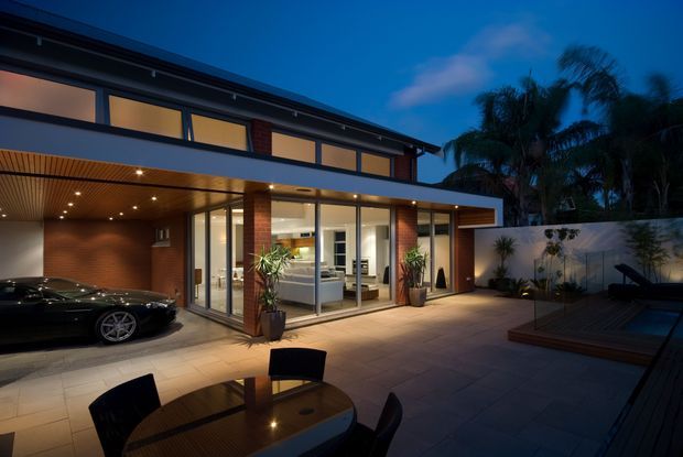 Dusk exterior view of a contemporary multi-level residence opening onto a paved courtyard and outdoor entertaining area. The home features broad floor-to-ceiling glazing, a projecting flat roof, exposed red brick walls, dark metal window frames, and a timber-lined soffit with recessed downlights. A carport with a black vehicle, outdoor dining setting, landscaped boundary, and partially visible swimming pool are present. The glazed living area is visible inside.