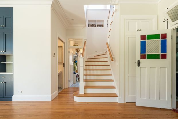 Bright traditional-style interior hallway with a central timber staircase leading to an upper landing and skylight. The space has polished warm timber flooring, white painted walls with decorative crown moulding, built-in blue-grey cabinetry on the left, and white paneled doors. A colourful geometric stained-glass-style panel is mounted on the open door at right.