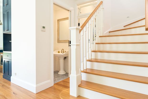 Bright traditional-style interior featuring a timber staircase with white painted risers, turned balusters, and a polished wood handrail. A small bathroom or powder room is visible beneath and beside the stairs, with a pedestal basin, patterned floor tile, and classic fittings. Built-in blue cabinetry and warm timber flooring are visible at the left.