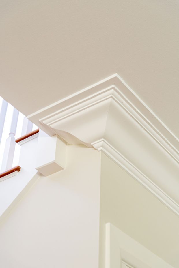 Close-up interior architectural detail showing ornate layered crown moulding and cornice trim around a ceiling and wall junction. The space is predominantly painted off-white/cream, with a glimpse of a bright stairwell or landing and white balustrade with dark wood handrails at left.
