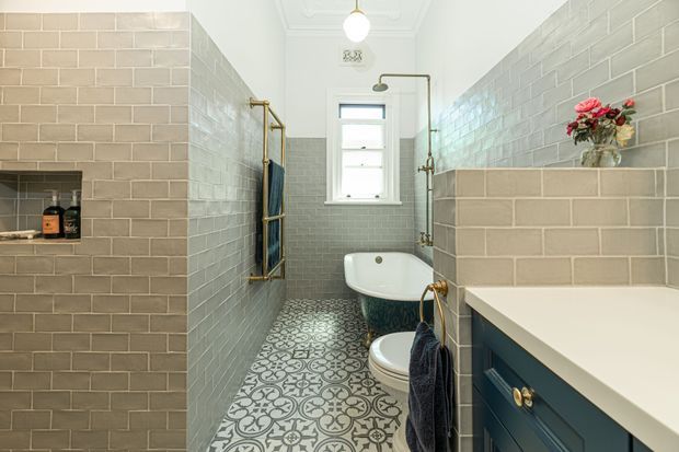 A narrow, bright traditional bathroom with full-height pale grey subway-tile walls, patterned encaustic-style floor tiles, a freestanding clawfoot-style bathtub, toilet, and a dark blue vanity with a light stone benchtop. Brass fixtures, a small high window, towel rails, and a floral arrangement add refined period-inspired detail.