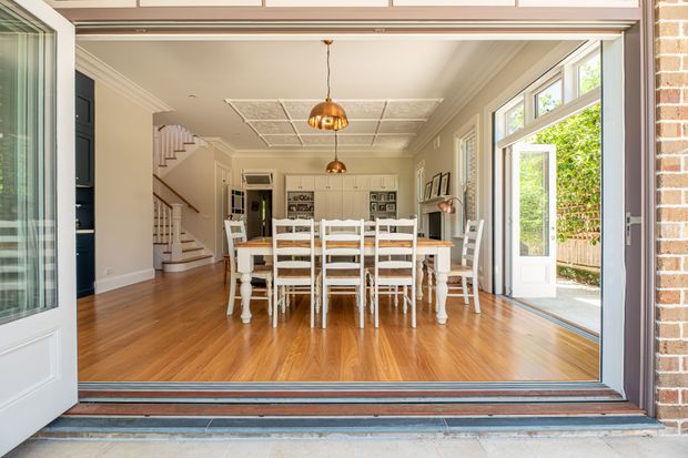 Bright traditional dining area viewed from an open exterior doorway, with polished timber flooring, a long white dining table and chairs, ornate ceiling detailing, pendant lights, large glazed doors, and a staircase leading to the upper level. Exposed brick is visible at the exterior edge.