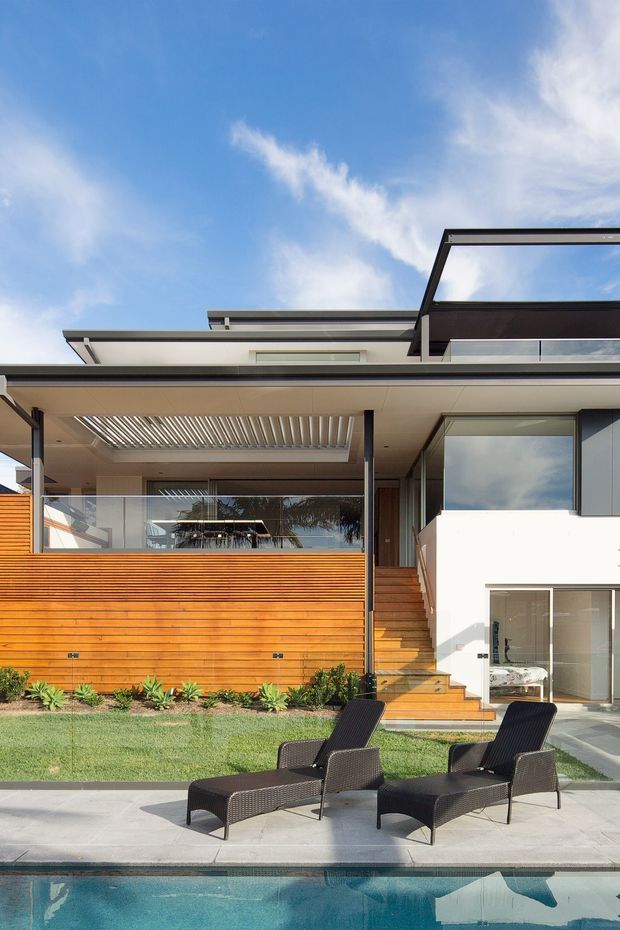 Exterior view of a contemporary multi-level residence with a flat roof, expansive glazing, timber-clad walls, glass balustrades, and a covered outdoor entertaining area. A landscaped lawn, poolside paving, and two outdoor lounge chairs occupy the foreground.