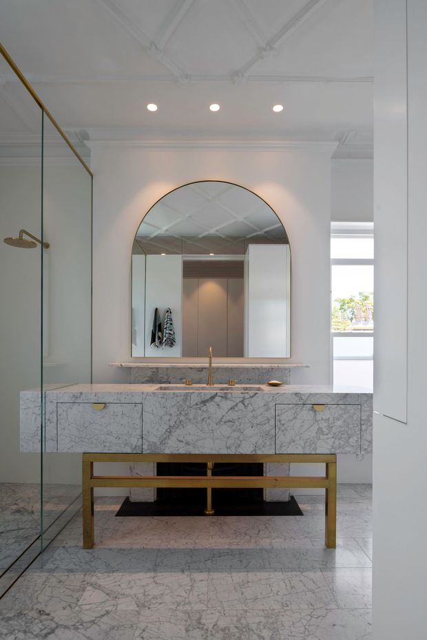 A refined, light-filled bathroom featuring a large marble double vanity with integrated drawers, brass tapware and a brass open support frame. A full-height frameless glass shower is visible on the left, while an arched brass-framed mirror sits above the vanity. The room has extensive white and grey veined marble surfaces, ornate decorative ceiling detailing, recessed lighting, and a bright window beyond.