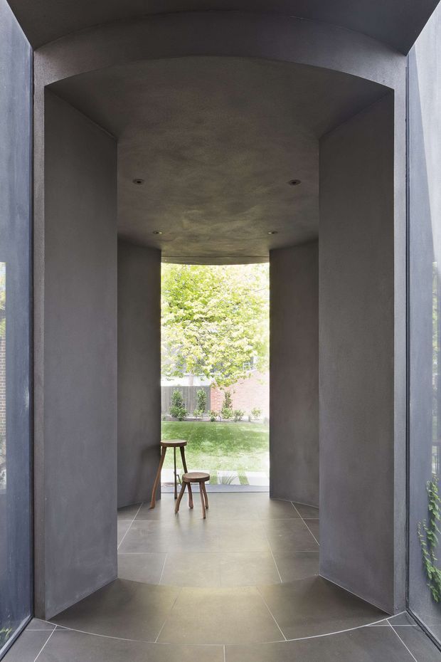 A dramatic, narrow architectural passage or entry vestibule with dark grey plastered walls and a curved, vaulted ceiling. Large glazed openings at the sides and rear frame a leafy garden courtyard. The floor is finished in large-format grey tiles, with two small wooden stools placed near the garden-facing opening. Recessed downlights and sharply geometric wall planes create a minimalist, sculptural composition.