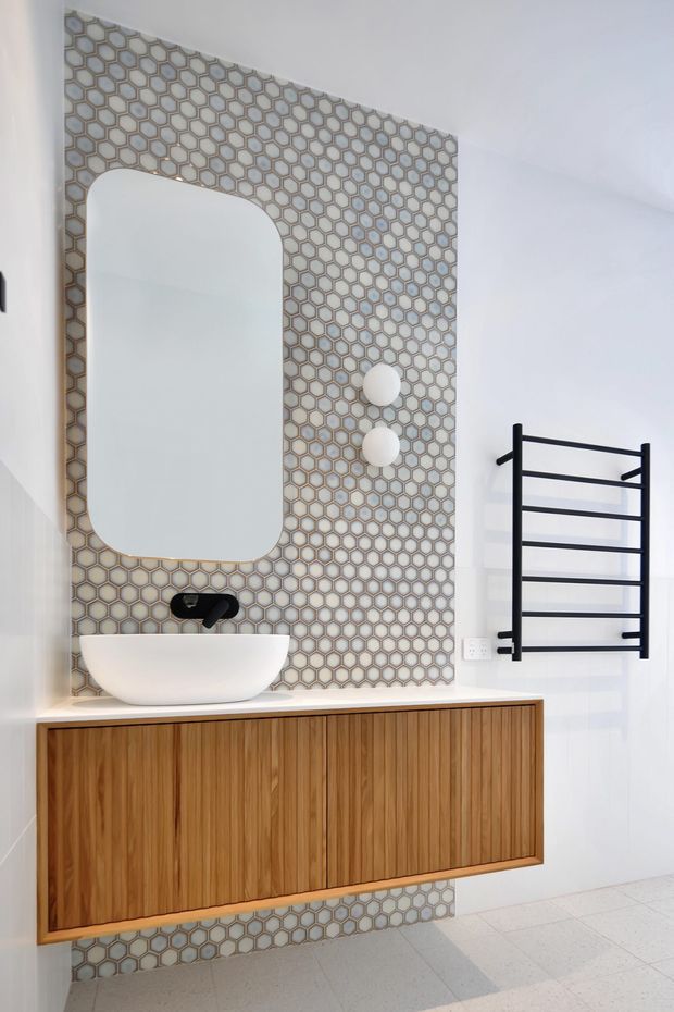 Contemporary bathroom vanity featuring a floating timber cabinet with vertical slatted fronts, a white vessel basin, black wall-mounted tapware, and a rounded-rectangle mirror. A geometric honeycomb tile splashback in soft grey and beige tones extends behind the vanity, complemented by a black heated towel rail and pale tiled flooring.