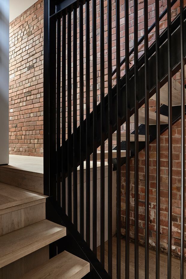Close-up interior view of a contemporary stairway with light timber treads, a black metal balustrade of closely spaced vertical bars, and exposed red brick walls. The stair structure and industrial material palette dominate the frame.