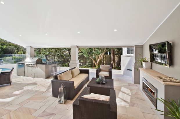 Bright covered outdoor entertaining area with a contemporary living room arrangement, outdoor kitchen and built-in barbecue, dining and lounge furniture, tiled flooring, and large openings to a landscaped garden. A wall-mounted television and linear fireplace create an outdoor media and relaxation zone.