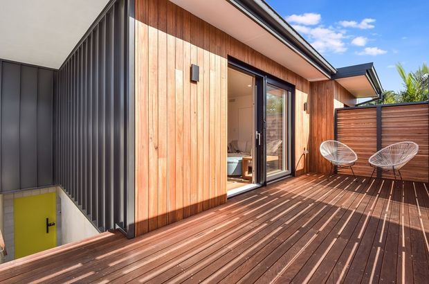 Contemporary timber-clad exterior with a broad raised timber deck and privacy screening. The image shows vertical warm timber wall cladding, dark corrugated metal cladding, dark-framed glazing and a compact outdoor seating area. A bedroom is visible through the open glazed door, while the deck overlooks a lower-level stair access. Bright sunny setting with clean modern detailing.