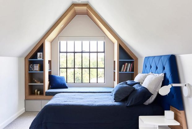 A bright attic bedroom with a steeply pitched gable ceiling and exposed timber trim framing a large black-grid window. Built-in dark timber shelving and a window seat flank the glazing, while the room features a deep blue upholstered bedhead, navy bedding, white walls, and a small white bedside table with a modern wall lamp.