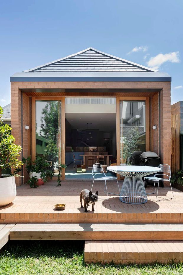 A sunny rear courtyard view of a brick home with a raised brick patio and timber-framed glazed opening leading into the interior. The patio contains a round white wire-frame table, matching outdoor chairs, potted plants, and a barbecue, with a small dog in the foreground. A hipped tiled roof and clear blue sky are visible above.