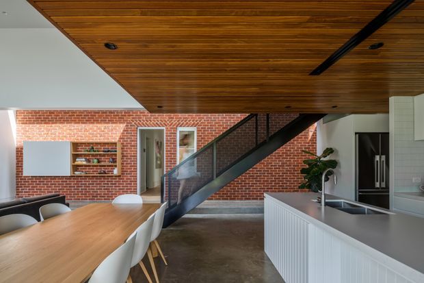 Open-plan kitchen and dining area with a dramatic black metal stair rising across the room. The space features exposed red brick walls, a warm timber-lined ceiling, polished concrete flooring, a large timber dining table with white chairs, and a white kitchen island with integrated sink and dark appliances. The overall design combines strong industrial architectural elements with clean contemporary joinery.