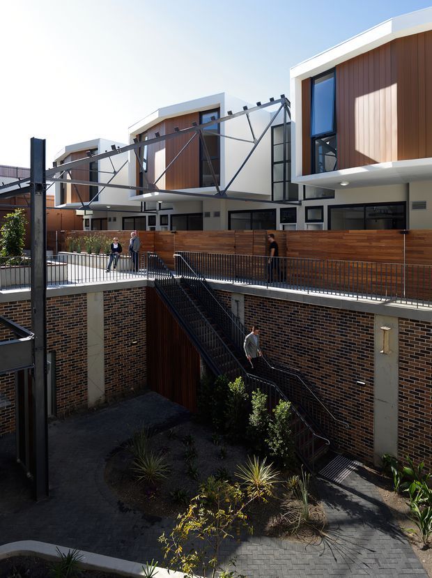 Exterior view of a contemporary multi-level residential or mixed-use building arranged around a sunken landscaped courtyard. The upper facade uses warm vertical timber cladding with crisp white projecting frames, dark-framed windows and extensive open steel bracing or structural supports. A timber privacy fence, brick retaining walls, metal balustrades and a stairway connect the upper walkway to the lower courtyard. Bright daylight and strong geometric lines define the scene.